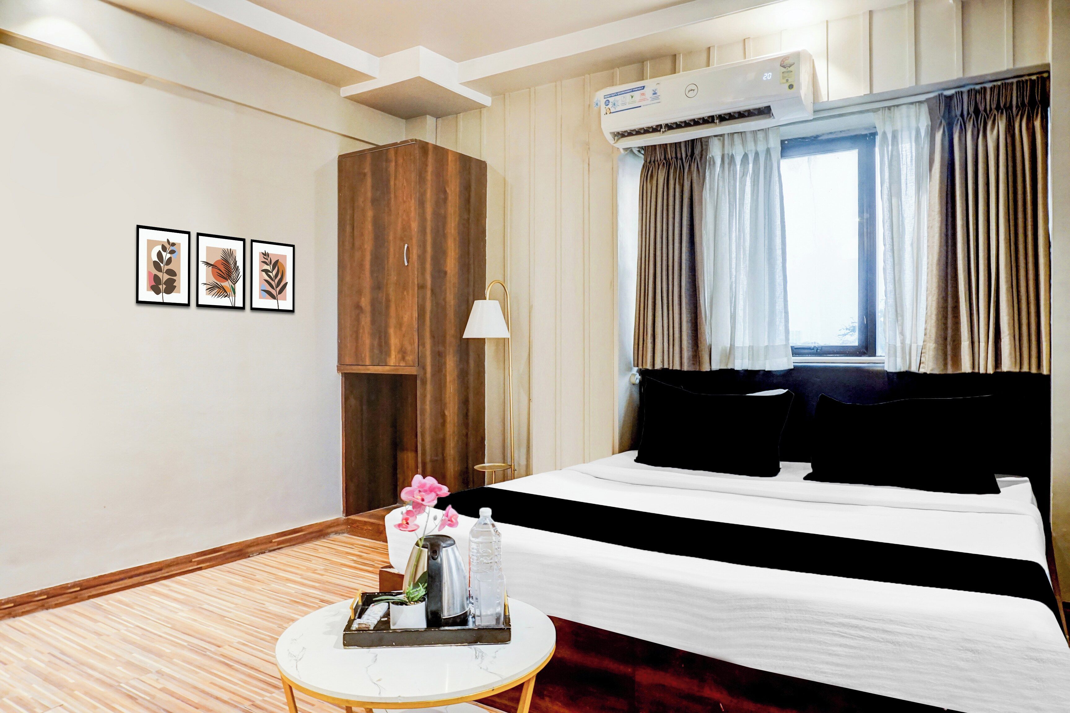 A hotel room with a bed, a bedside table with a tray, a wall-mounted air conditioner, and a window with curtains.