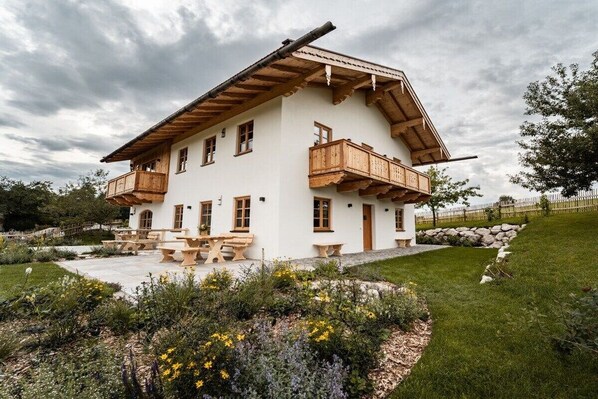A two-story chalet with a wooden balcony, a stone wall, and a garden with flowers. - Home - Apartment Liesl (Surberg)