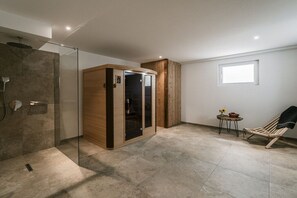 A modern bathroom with a glass-enclosed shower, a wooden cabinet, and a small table with a chair. - Home - Apartment Liesl (Surberg)