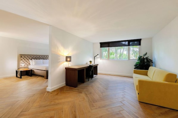 A modern room with a herringbone floor, a bed with a tufted headboard, a desk with a lamp, and a yellow sofa. - Parasasa Hotel Curacao (Willemstad)