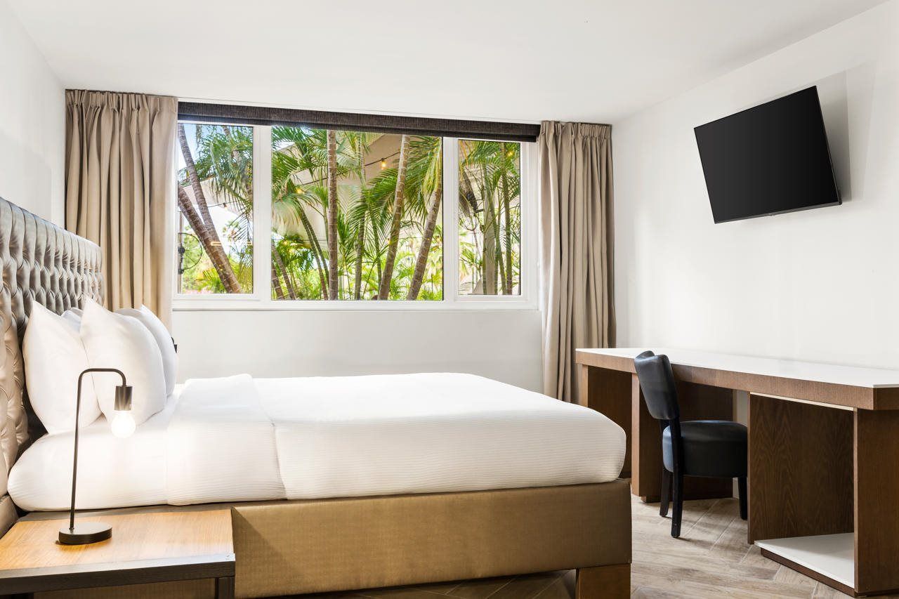Una habitación de hotel con cama, escritorio, silla, televisor y ventana con cortinas.