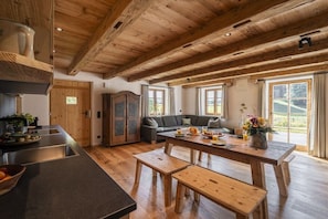A spacious kitchen with wooden beams, a dining area with a wooden table and benches, and a view of the outdoors through large windows. - Home - Apartment Ludwig (Surberg)