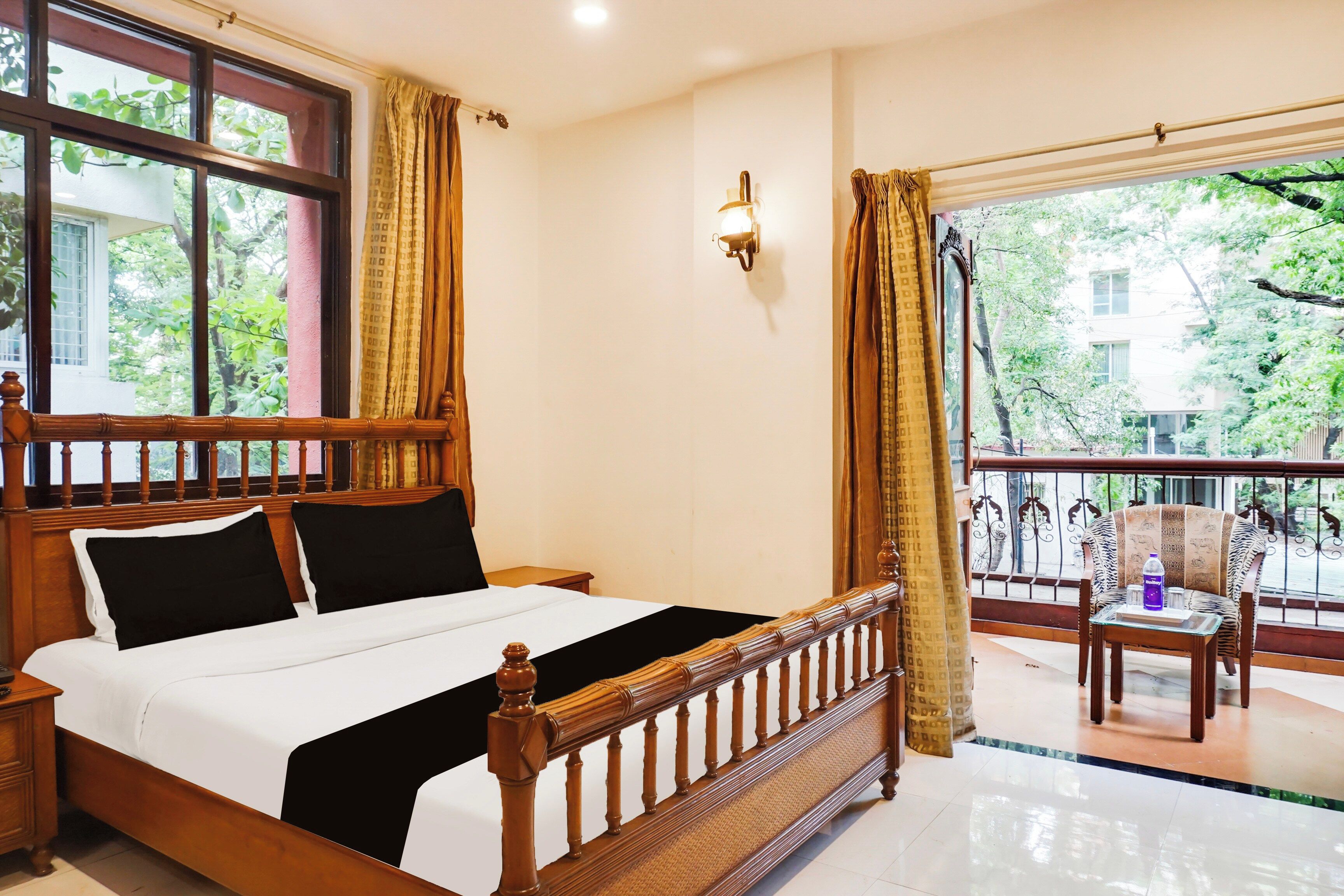 A hotel room with a bed, a nightstand, a window with curtains, a wall-mounted light, and a balcony with a chair and table.