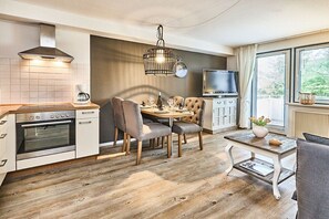 A modern kitchen with a dining area, wooden flooring, a built-in oven, a coffee machine, a TV, and a window with a view. - Holiday apartment with 1 bedroom (Westerland)