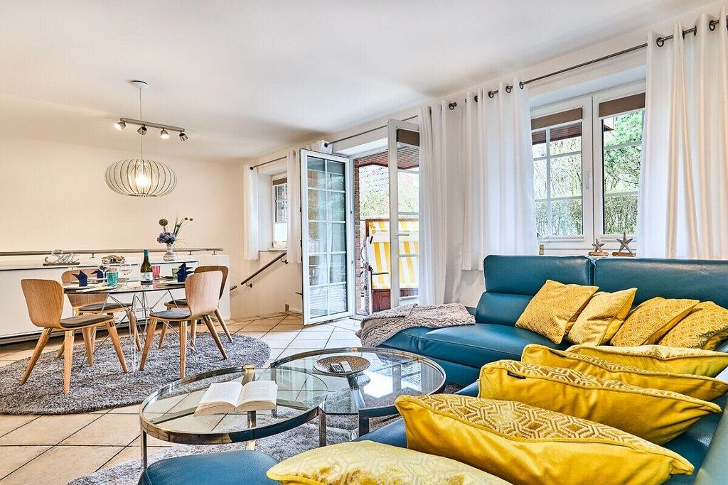 Un salon avec un canapé bleu, des coussins jaunes, une table basse en verre et un coin repas comprenant des chaises et une table.