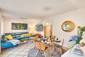 Ein modernes Wohnzimmer mit einem blauen Sofa, einem Esstisch, Stühlen, einem farbenfrohen abstrakten Gemälde und einem dekorativen Spiegel.