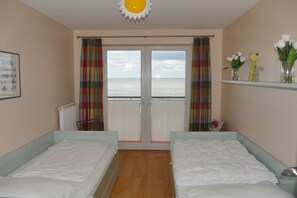 Een kamer met twee bedden, uitzicht op de zee door een deur en een plank met bloemen.
