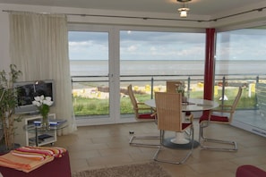 A modern living room with a large window overlooking a beach, a television, and a dining area with chairs. - Strandpalais, Apt. 508 (Cuxhaven)