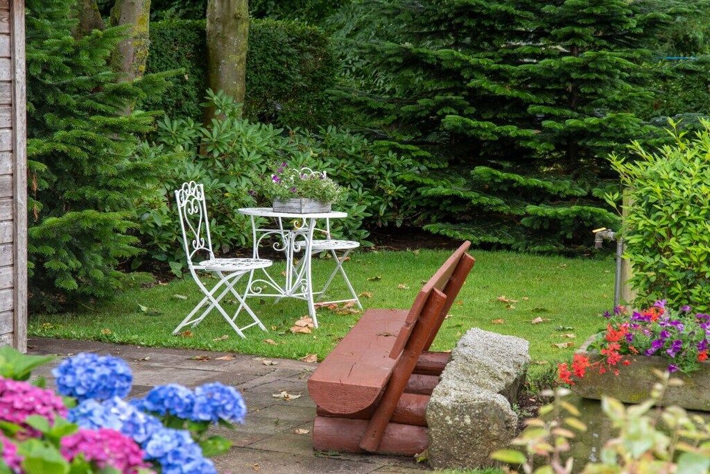 A garden with a white metal table and chairs, surrounded by lush greenery and a wooden bench.