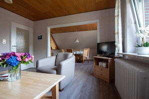 A living room with a sofa, a TV, a table with a vase of flowers, and a dining area in the background. - Siewertsen Apt. No. 1 (Alkersum)