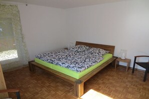 Ein Zimmer mit einem Einzelbett, einem hölzernen Bettgestell, einem gemusterten Lattenrost, einem kleinen Nachttisch, einem Stuhl und einem Fenster mit Vorhang.