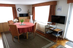 Ein Hotelzimmer mit einem runden Esstisch, Stühlen, einem Flachbildfernseher, einer kleinen Kommode, einer Uhr und einem Fenster mit Vorhängen.