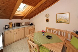 Cocina compacta con armarios de madera, microondas y cafetera. Hay una mesa de comedor con mantel verde y un jarrón de flores.