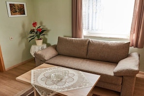 A living room with a beige sofa, a small table with a doily, a potted plant, and a window with curtains. - Holiday apartment with 1 bedroom (Röslau)
