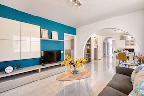 A modern living room with a flat-screen TV, a clock, and a coffee table with flowers. - Spacious & bright apartment, sun terrace PGAL1-1 (Sliema)