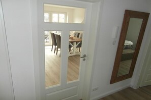 Une porte blanche avec un panneau de verre, un miroir encadré de bois, et un coin repas bien visible avec des chaises et une table à l’arrière-plan.