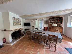A dining area with a long table, chairs, a fireplace, a television, and a cabinet with glassware.
