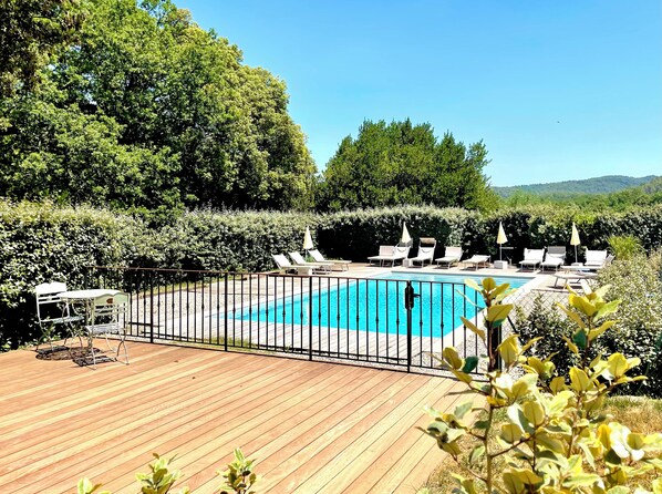 A wooden deck with a pool, surrounded by lounge chairs and umbrellas, set against a backdrop of trees and hills.
