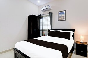 Una habitación de hotel con cama, mesita de noche con lámpara, unidad de aire acondicionado, ventana con cortinas y un cuadro en la pared.