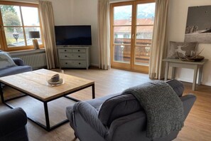 A modern living room with a sofa, a coffee table, a television, and a view through large windows. - Holiday apartment with 1 bedroom (Garmisch-Partenkirchen)