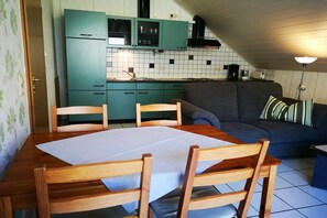 Una cocina compacta con armarios verde azulado, una mesa de comedor con sillas y un sofá gris con cojines a rayas.