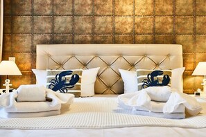 Quarto de hotel com cabeceira estofada, duas luminárias de apoio e almofadas decorativas com desenho de caranguejo.