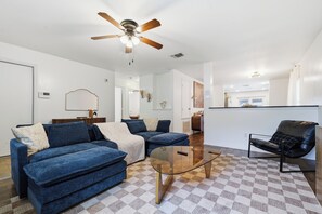 Ein modernes Wohnzimmer mit Deckenventilator, einem Glastisch und einer blauen Sofagarnitur.