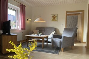Ein Wohnzimmer mit Sofa, Sesseln, Couchtisch und Fernseher auf einem Holzkasten.