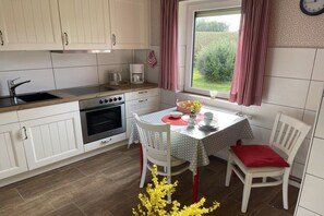 A compact kitchen with white cabinets, a stove, and a sink. There's a dining table with chairs and a window with curtains. - "Mullewap" at the Koch holiday farm (Schönhagen)