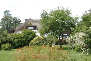 A traditional thatched-roof house surrounded by a lush garden with various shrubs and flowering plants. - "Mullewap" at the Koch holiday farm (Schönhagen)