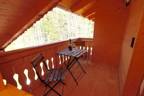 Eine Holzterrasse mit Tisch und Stühlen, einer Weinflasche und Blick auf Bäume.