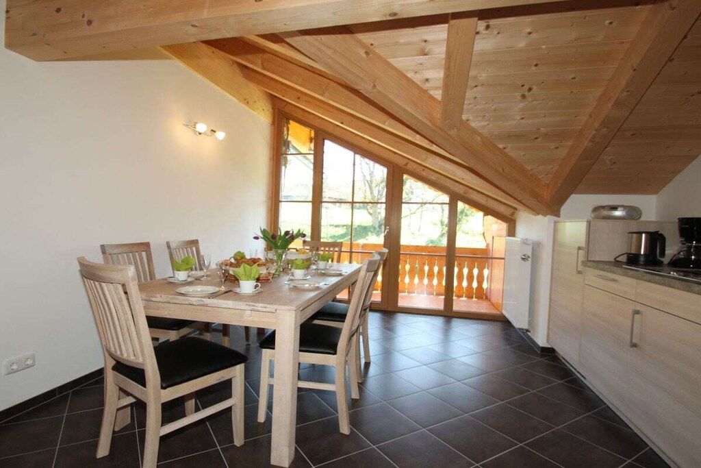 A dining area with a wooden table and chairs, a kitchen with a refrigerator and stove, and a large window with a view of greenery.