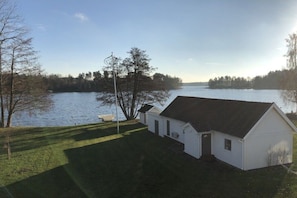 A white building with a dark roof, situated next to a body of water, surrounded by trees and a grassy area. - Holiday home 25 m from Lake Nömmen (Stensjön)
