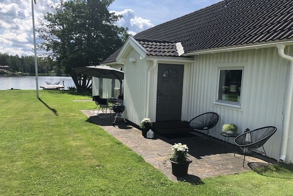 A white building with a dark roof, a covered patio area with chairs, and a view of a lake. - Holiday home 25 m from Lake Nömmen (Stensjön)