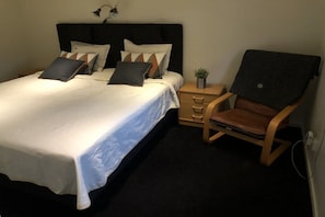 A neatly made bed with a white comforter and multiple pillows, a wooden nightstand with a small plant, and a brown leather chair. - Holiday home 25 m from Lake Nömmen (Stensjön)