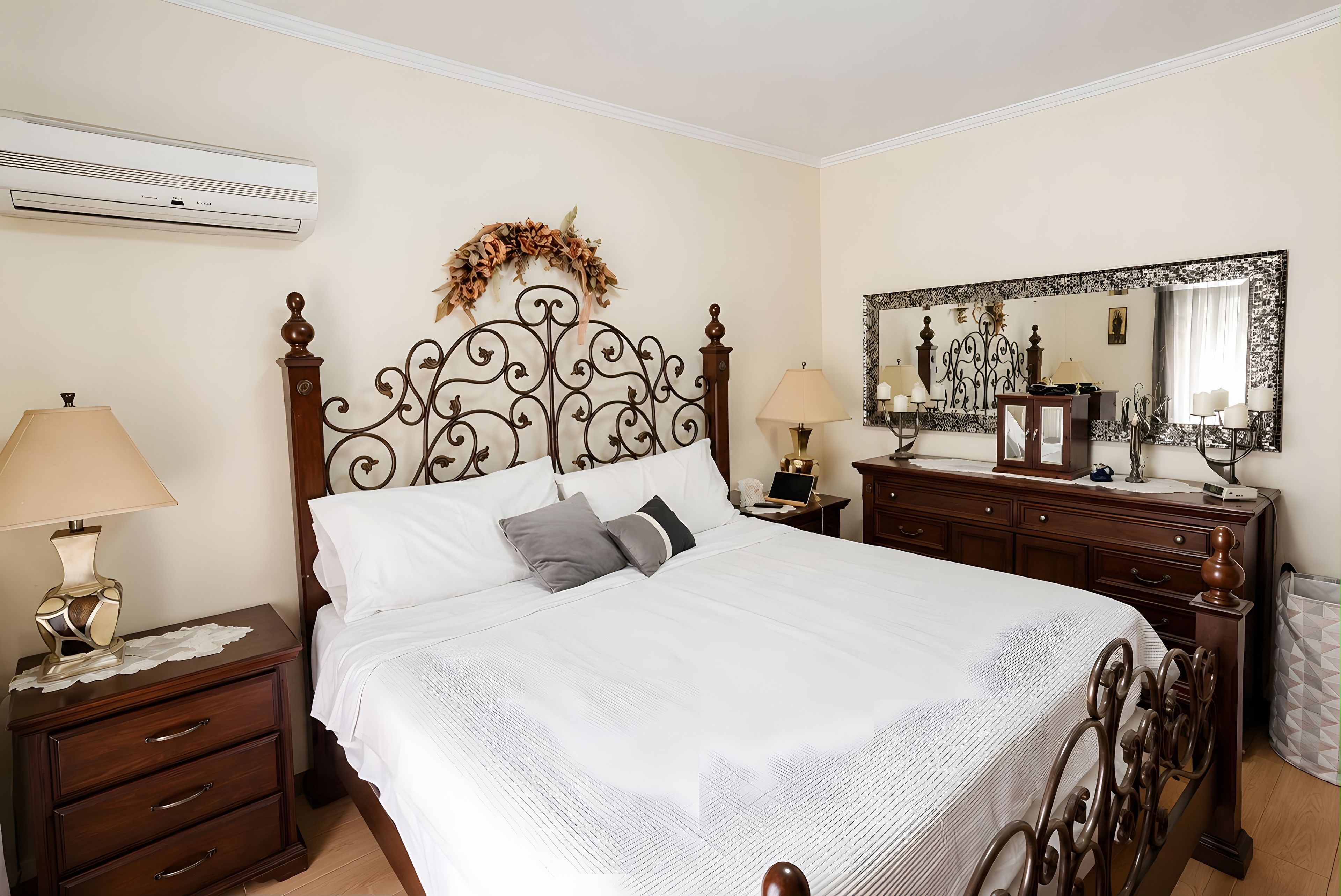 Een net opgemaakt bed met een decoratief hoofdbord, nachtkastjes met lampen, een dressoir met spiegel en een wandairconditioner.