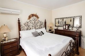 A neatly made bed with a decorative headboard, bedside tables with lamps, a dresser with a mirror, and a wall-mounted air conditioner.
