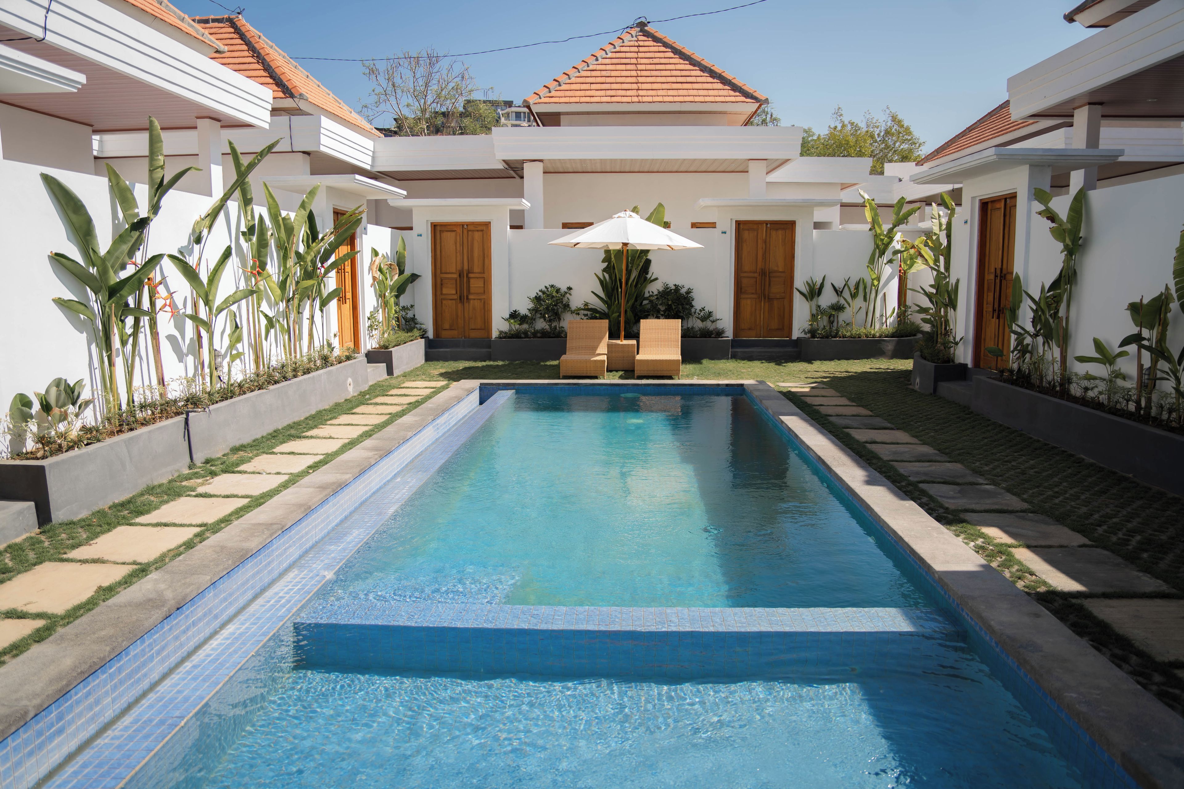 A rectangular swimming pool with a tiled edge, surrounded by a garden with various plants and a seating area with chairs and an umbrella.