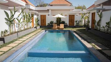 A rectangular swimming pool with a tiled edge, surrounded by a garden with various plants and a seating area with chairs and an umbrella.