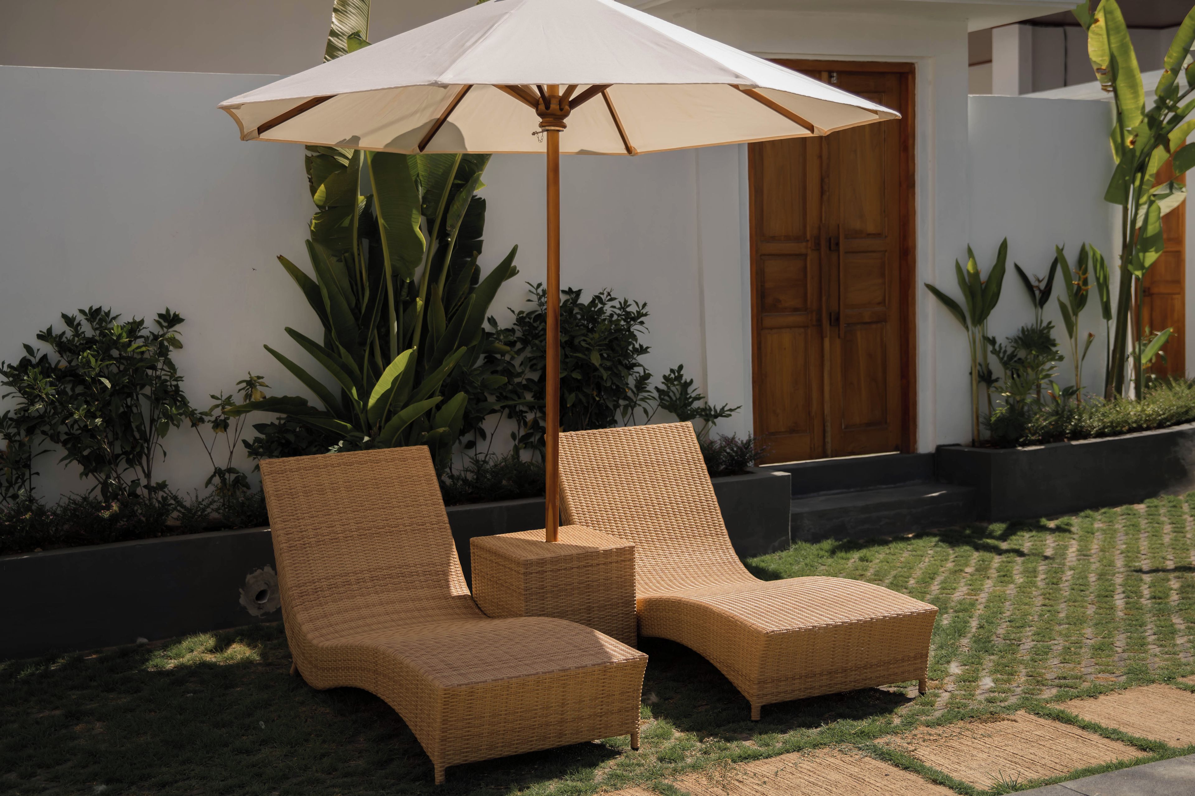 Two wicker lounge chairs with a canopy umbrella, positioned on a grassy area with a white wall and wooden door in the background.