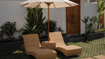 Two wicker lounge chairs with a canopy umbrella, positioned on a grassy area with a white wall and wooden door in the background.