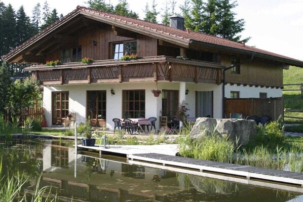 Uma casa de madeira de dois andares com varanda, lago e mobiliário de exterior.
