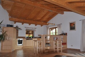 A spacious kitchen with wooden beams, a dining area with chairs, and a view of the outdoors through a window. - Holiday apartment with 1 bedroom (Rieden am Forggensee)