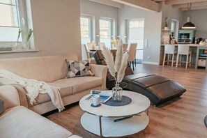 A modern living room with a sofa, a coffee table, and a dining area in the background. - Holiday home with 1 bedroom (Maasholm-Bad)