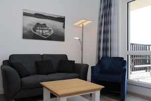 A modern living room with a grey sofa, a blue armchair, a wooden coffee table, a floor lamp, and a large window with a view of a balcony. - Apartment 15 in Villa Meeresblau (Cuxhaven)