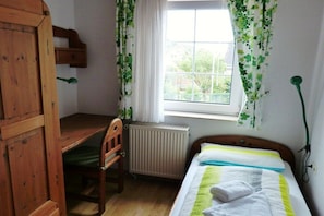 Una habitación con escritorio de madera, una silla, cama con colcha a rayas blancas y verdes y ventana con cortinas blancas.
