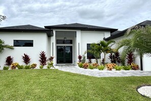 A modern two-story house with a flat roof, a front entrance with a glass door, and a well-maintained lawn with various plants. - Villa in Cape Coral with swimming pool (Cape Coral)