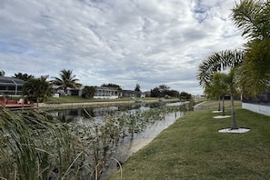 A calm waterway with a grassy bank, palm trees, and a building with a sloped roof in the background. - Villa in Cape Coral with swimming pool (Cape Coral)