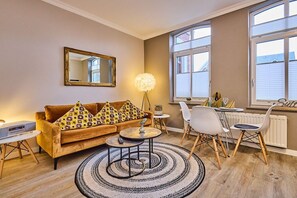 A furnished living room with a sofa, two side tables, a lamp, and a dining area with chairs and a table. - Holiday apartment with 1 bedroom (Westerland)