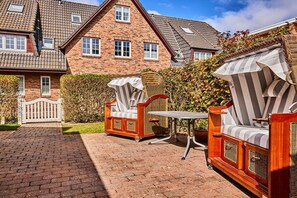 Ein Ziegelhaus mit gepflasterter Auffahrt und zwei Außenbereichen zum Sitzen, ausgestattet mit Korbmöbeln und gestreiften Kissen.